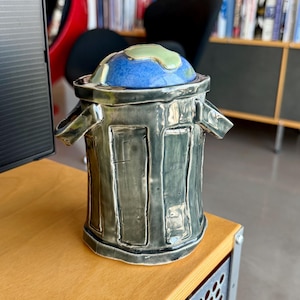 May include: A ceramic trash can-shaped container with a lid. The lid is blue with a green and gold continents design. The body of the container is gray with rectangular panels and handles. The container is sitting on a wooden surface.