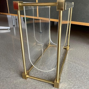 Könnte beinhalten: Ein Zeitschriftenständer aus klarem Acryl und Messing. Der Ständer hat einen rechteckigen Messingrahmen mit vier vertikalen Messingstützen und transparenten Acrylplatten. Das Messing hat eine polierte Oberfläche.