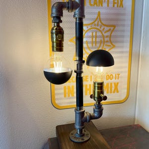 May include: A table lamp constructed from dark grey industrial metal pipes and fittings mounted on a rectangular wooden base. The lamp features two brass light sockets holding clear globe bulbs, each topped with a matte black hemispherical shade.