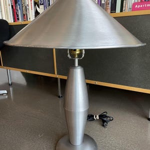 Könnte beinhalten: Eine silberfarbene Metall-Tischlampe mit konischem Schirm und rundem Fuß. Die Lampe hat ein modernes Design mit gebürsteter Metalloberfläche. Die Lampe ist eingesteckt. Die Lampe steht auf einem grauen Boden. Die Lampe ist ca. 60 cm hoch.