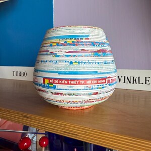 May include: A decorative, handcrafted vase with a unique design. The vase is made from layered paper strips, featuring text and colorful patterns. The vase has a rounded shape and sits on a wooden shelf. The text includes "XỔ SỐ KIẾN THIẾT TP. HỒ CH&Iacute; MINH".