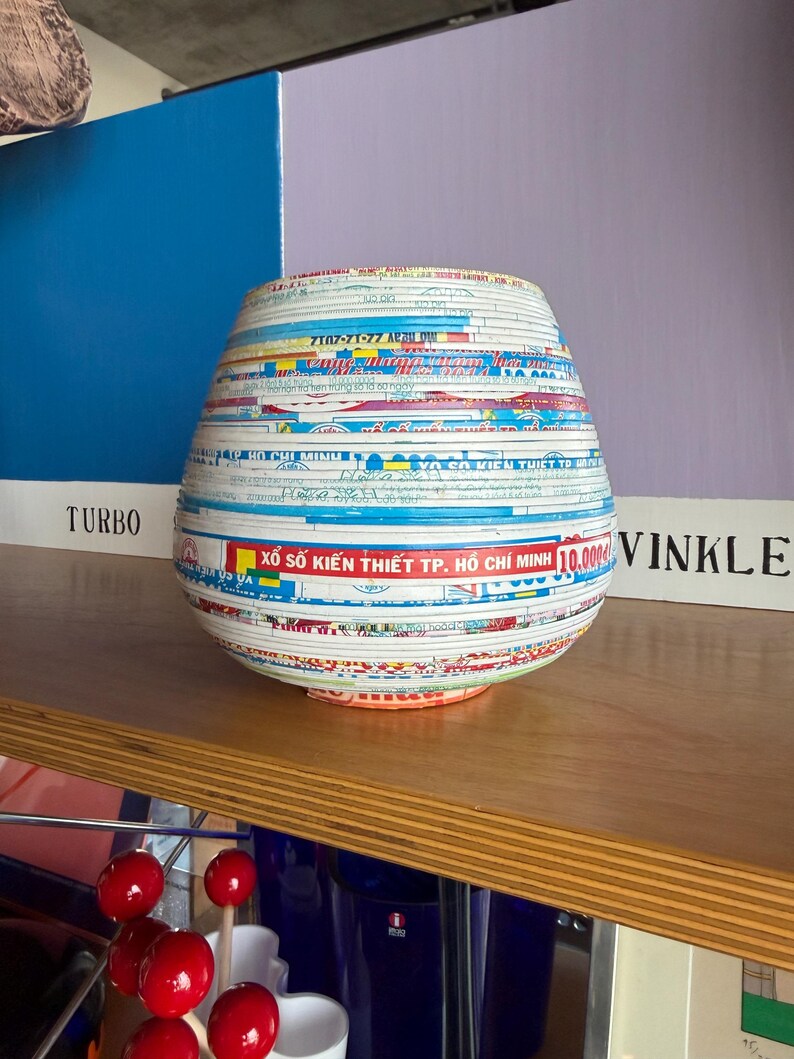 May include: A decorative, handcrafted vase with a unique design. The vase is made from layered paper strips, featuring text and colorful patterns. The vase has a rounded shape and sits on a wooden shelf. The text includes "XỔ SỐ KIẾN THIẾT TP. HỒ CH&Iacute; MINH".