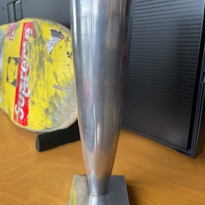 May include: A silver metal vase with a tall, narrow, conical shape and a square base. The vase is sitting on a wooden surface.