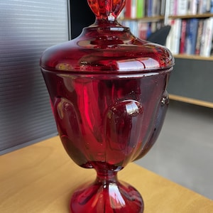 Może przedstawiać: Rubinowoczerwony szklany pojemnik na cukierki z pokrywką. Naczynie ma podstawę i pękaty korpus z teksturowanym wzorem. Pokrywka zwieńczona jest ozdobnym zwieńczeniem. Pojemnik na cukierki jest prawdopodobnie w stylu vintage.