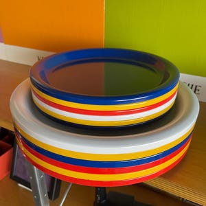 Może przedstawiać: Stos kolorowych, okrągłych talerzy z czerwonymi, żółtymi, niebieskimi i białymi paskami. Talerze są wykonane z plastiku i idealnie nadają się do serwowania przekąsek lub deserów.