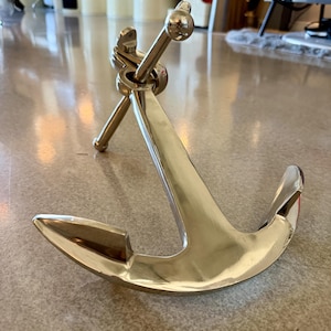 May include: A polished brass anchor with a curved design. The anchor has a shiny, reflective surface and is designed with a pointed fluke and a rounded ring at the top. The anchor is a nautical decorative item.