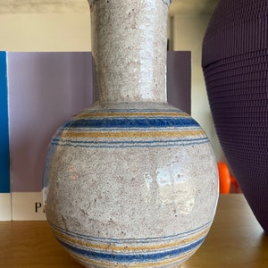 以下が含まれることがあります： 青と黄色のストライプが入った白い陶器の花瓶。花瓶は丸い胴体と背が高く細い首をしています。