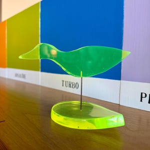 May include: A neon green acrylic bird sculpture with a matching base. The bird is mounted on a thin metal rod. The word "TURBO" is visible in the background. The sculpture is displayed on a wooden surface.