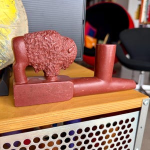 Puede incluir: Una pipa de piedra tallada de color marrón rojizo con una figura de bisonte. La pipa tiene una base rectangular y un cuenco cilíndrico. El bisonte está detallado con pelaje texturizado. La pipa se exhibe sobre una superficie de madera.