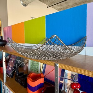 May include: A square, silver-toned metal wire mesh bowl with curved, raised edges. The bowl features a woven grid pattern and is positioned on a wooden shelf, showcasing a metallic finish and a structured, open-weave design throughout the piece.