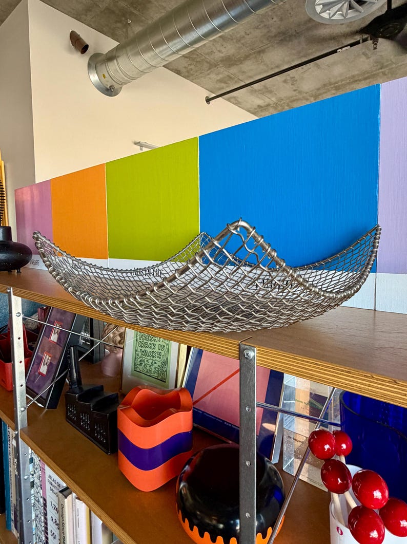 May include: A square, silver-toned metal wire mesh bowl with curved, raised edges. The bowl features a woven grid pattern and is positioned on a wooden shelf, showcasing a metallic finish and a structured, open-weave design throughout the piece.