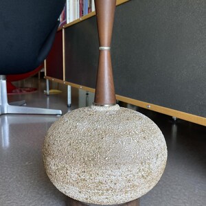 May include: A mid-century modern table lamp with a textured, spherical base in beige and brown tones. The lamp features a wooden neck and base, and a brass-colored lamp holder. The lamp is approximately 24 inches tall.