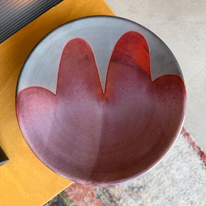 Puede incluir: Un plato redondo de cerámica con borde gris y diseño central en tonos rojos y morados. El diseño se asemeja a dos formas de flores estilizadas. El plato está sobre una superficie amarilla.