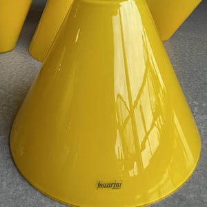 May include: Several bright yellow, cone-shaped glass lampshades. The lampshades have a glossy finish and a wide base that tapers to a narrow top. The brand name "foscarini" is printed on the side of one lampshade.