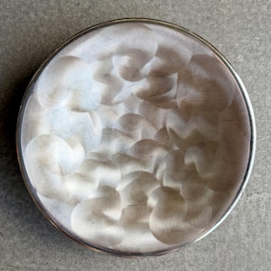 Puede incluir: Un plato redondo de metal plateado con un patrón arremolinado texturizado. El plato tiene un borde elevado y un acabado de metal cepillado. El diseño crea un aspecto artístico único. Adecuado para servir o exhibir.