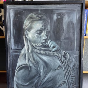 May include: A monochrome painting of a person with long hair, wearing a scarf and holding a plant. The artwork is framed in a black wooden frame. The painting is displayed against a backdrop of books.