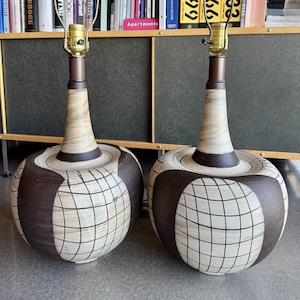 Puede incluir: Dos lámparas de mesa a juego con una base en forma de globo. La base es de color crema con un patrón de cuadrícula marrón oscuro y detalles. Las lámparas tienen un cuello marrón y un portalámparas de latón. Las lámparas están sobre una superficie gris.