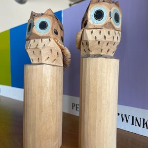 Könnte beinhalten: Zwei Holz-Eulen-Skulpturen mit hellbraunen Körpern, dunkelbraunen Flecken und blauen Augen. Die Eulen stehen auf zylindrischen Holzsockeln. Der Hintergrund umfasst eine lila Wand und einen farbenfrohen Abschnitt.