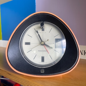 May include: A vintage Panasonic alarm clock with a triangular shape. The clock has a black face with white numbers and hands, and a red second hand. The clock is encased in a black and orange frame. The clock face reads "PANASONIC".