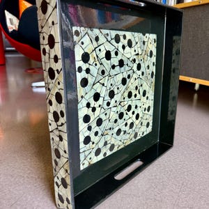 May include: A square black serving tray featuring a central panel with a cream-colored background, black circular dots, and a network of thin black crackle lines. The tray has integrated side handles and a glossy finish on the interior surface.