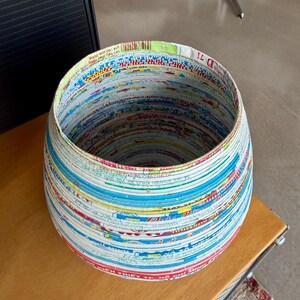 May include: A round, handcrafted basket made from recycled paper. The basket features a colorful, layered design with visible text and patterns. The interior is a dark space, and the basket sits on a wooden surface.