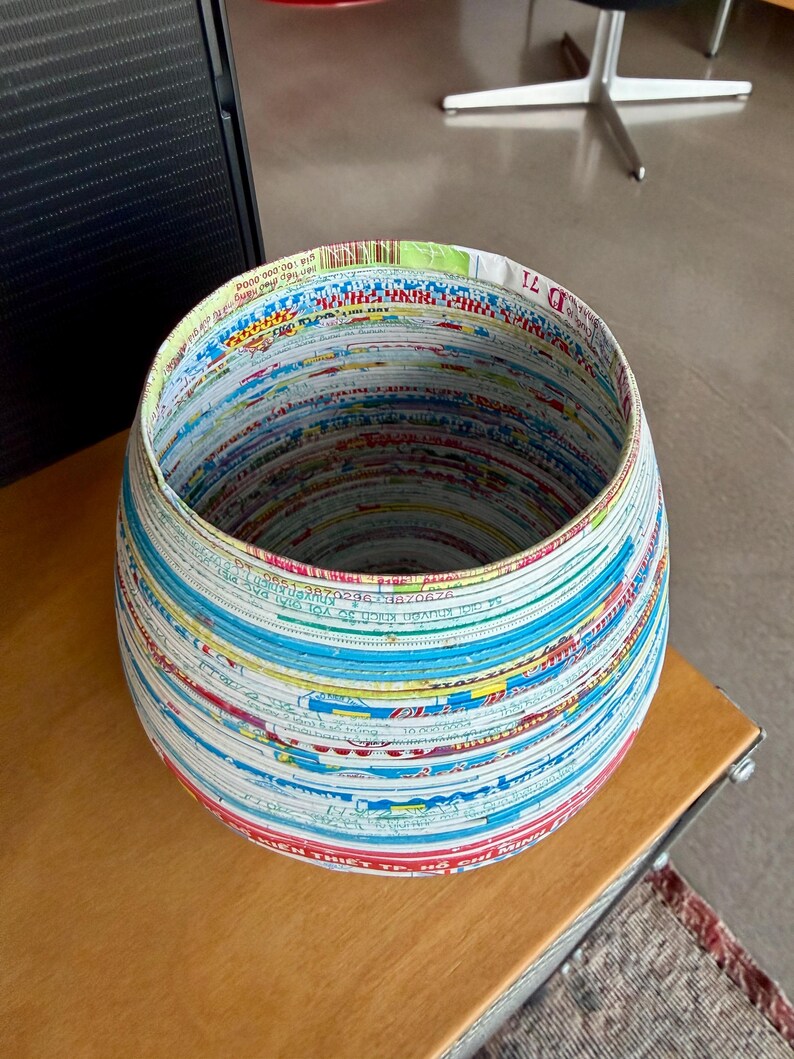 May include: A round, handcrafted basket made from recycled paper. The basket features a colorful, layered design with visible text and patterns. The interior is a dark space, and the basket sits on a wooden surface.