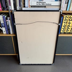 May include: The back of a framed picture with a black frame. The artwork is set against a cream-colored backing. A wire is attached for hanging. Bookshelves filled with books are visible in the background.