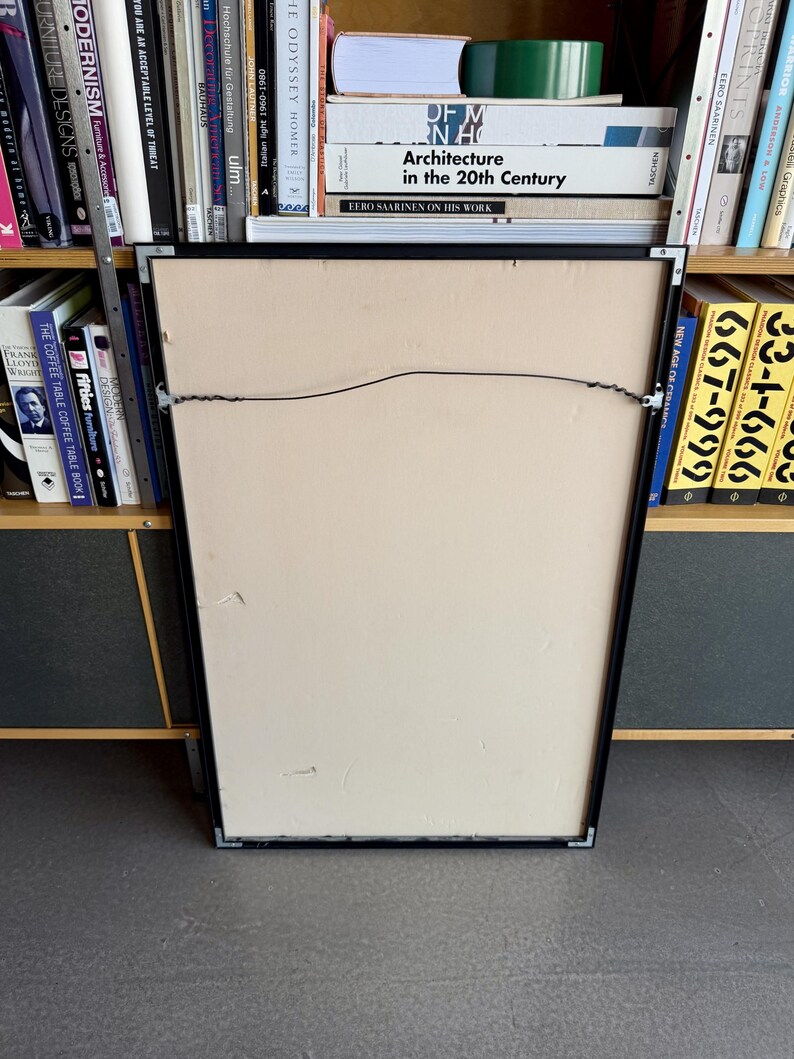 May include: The back of a framed picture with a black frame. The artwork is set against a cream-colored backing. A wire is attached for hanging. Bookshelves filled with books are visible in the background.