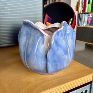 Puede incluir: Un jarrón de cerámica azul con forma de flor y un interior rosa claro. El jarrón tiene un diseño texturizado en forma de pétalo y está sobre una superficie de madera. Es un objeto decorativo.