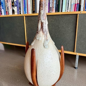 May include: A vintage ceramic table lamp with a speckled, off-white body and a brown, textured top. The lamp is supported by a wooden stand with three curved legs. The lamp is a unique piece of home decor.