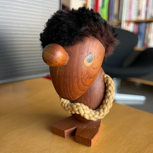 May include: A wooden figurine with a dark brown body, a light brown ear, and a black fur-like head. A rope-like detail encircles the neck. The figurine is standing on a wooden base. The eye is a silver-colored detail.