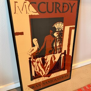 May include: Framed art print with the name "MCCURDY" in large letters. The artwork shows a person in a window with a bird, with the text "OBRAS SANJUANERAS" and "MCMXVIII" at the bottom. The color scheme includes beige, brown, and black.