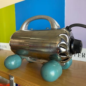 May include: A vintage silver-colored electric massager with a handle and four teal-colored wheels. The massager is on a wooden shelf with a blue, green, and purple background.