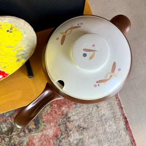 May include: A brown handled casserole dish with a white lid. The lid has a small handle and is decorated with brown, pink, and gray abstract designs. The dish sits on a patterned rug.