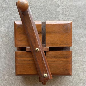 Peut inclure: Une boîte en bois avec un couvercle à charnière et une poignée en bois. La boîte est en bois brun foncé et comporte des vis en laiton. La poignée a une boule en bois à l'extrémité. La boîte est posée sur une surface grise.