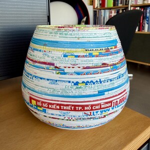 May include: A handcrafted, round vase made from layered, colorful paper strips. The vase features a wide, open top and a rounded base. The paper strips display text and patterns in various colors, creating a unique, textured surface.