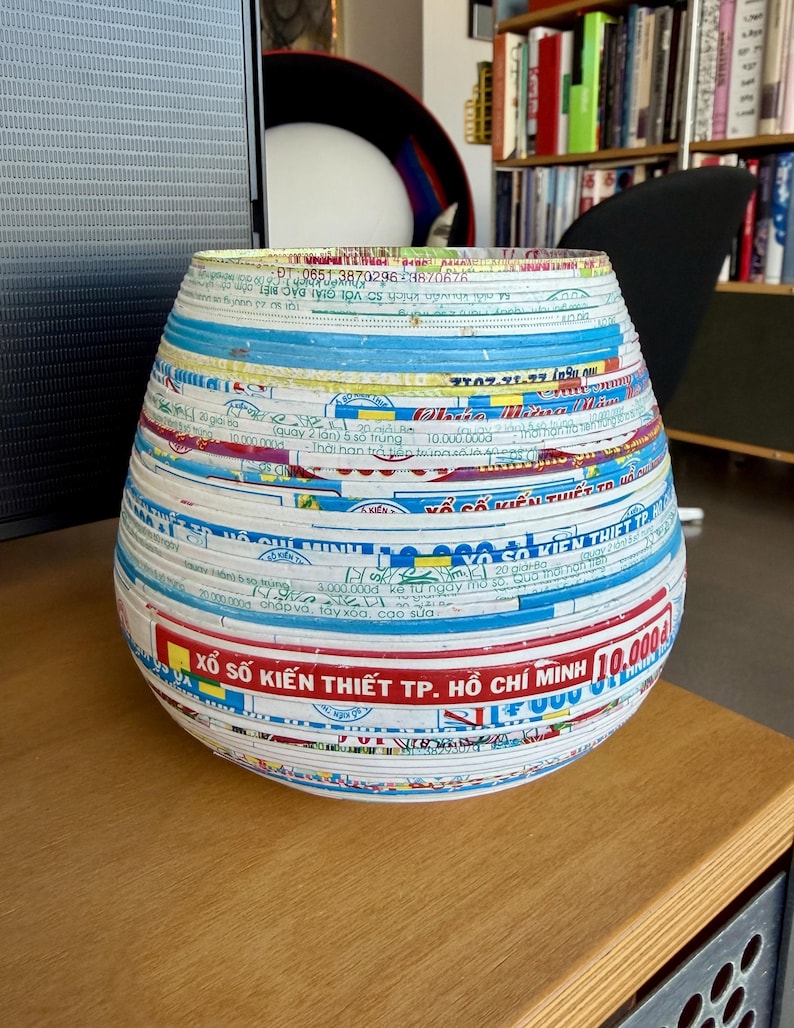 May include: A handcrafted, round vase made from layered, colorful paper strips. The vase features a wide, open top and a rounded base. The paper strips display text and patterns in various colors, creating a unique, textured surface.