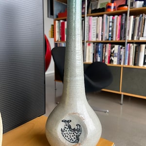 Può includere: Un vaso in ceramica alto e sottile con una smaltatura grigio chiaro e verde. Il vaso ha una base rotonda e un collo stretto. Un'illustrazione in bianco e nero di un pesce è dipinta sulla parte anteriore del vaso.
