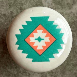 May include: A white ceramic knob with a teal and coral geometric design. The design features a diamond shape with a smaller diamond inside.