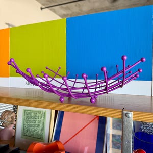 May include: A modern, purple metal fruit bowl with a unique, spiky design. The bowl sits on a wooden shelf, with colorful painted panels in the background. The bowl's design features a series of curved lines and spherical accents.