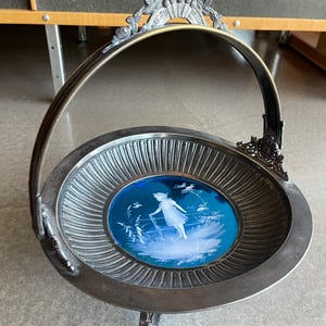May include: A silver serving dish with a blue glass insert featuring a white silhouette of a girl in a field. The dish has a handle and three legs.