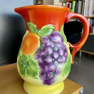 May include: A ceramic pitcher with a vibrant design. The top is a bright orange, transitioning to a body adorned with purple grapes, green leaves, and an orange fruit. The base is yellow, and the handle is red.