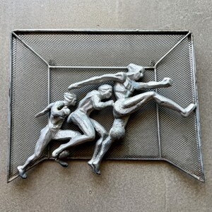May include: A silver-toned metal wall art piece featuring three stylized figures in motion, resembling soccer players in action. The figures are mounted against a rectangular wire mesh screen with a frame, creating a dimensional relief effect.