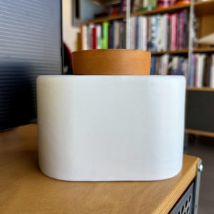 May include: A white, rectangular container with rounded edges, topped with a small, circular, light brown lid. The container sits on a wooden surface. The background includes a bookshelf and a computer monitor.