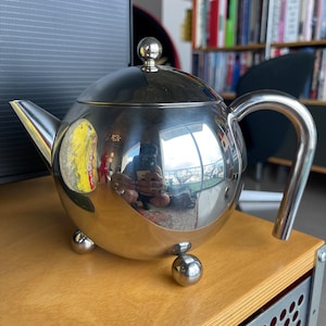 Puede incluir: Una tetera de plata brillante con cuerpo redondo y asa curva. La tetera tiene tres patas redondas y una tapa de plata brillante con un pomo en la parte superior.