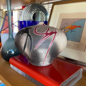 May include: A gray ceramic vase with black and red abstract designs sits on a red book with a black base. The vase is on a wooden shelf with other objects in the background.