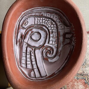 May include: A terracotta-colored, oval-shaped ceramic bowl with a raised, abstract design in white. The design features geometric shapes and swirling patterns, creating a unique decorative piece. The bowl's rim is a darker shade of brown.