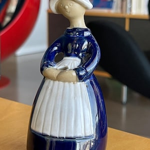 Puede incluir: Una figurita de cerámica de una mujer con un vestido azul y un delantal blanco. Lleva un sombrero blanco y tiene una cara sonriente.