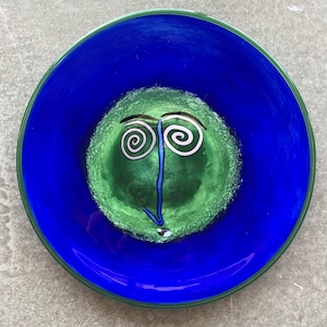 May include: A blue glass bowl with a green face design. The face has two white spiral eyes and a blue nose.