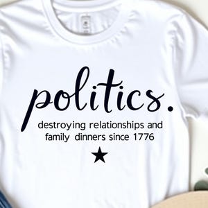 Op de afbeelding: Wit T-shirt met het woord "politics" in een cursief lettertype, samen met de zin "destroying relationships and family dinners since 1776" en een ster. Het shirt wordt gecombineerd met een blauwe jeans en een beige hoed.